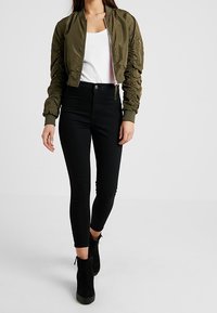 Kvinna som bär en beskuren, olivgrön bomberjacka, vit linne, högmidjade svarta skinny jeans och svarta ankelstövlar som går framåt.