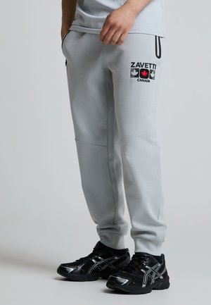 Person trägt hellgraue Jogginghose mit „Zavetti Canada“-Logo und schwarzen Sportschuhen mit weißen Akzenten, steht vor neutralem Hintergrund.