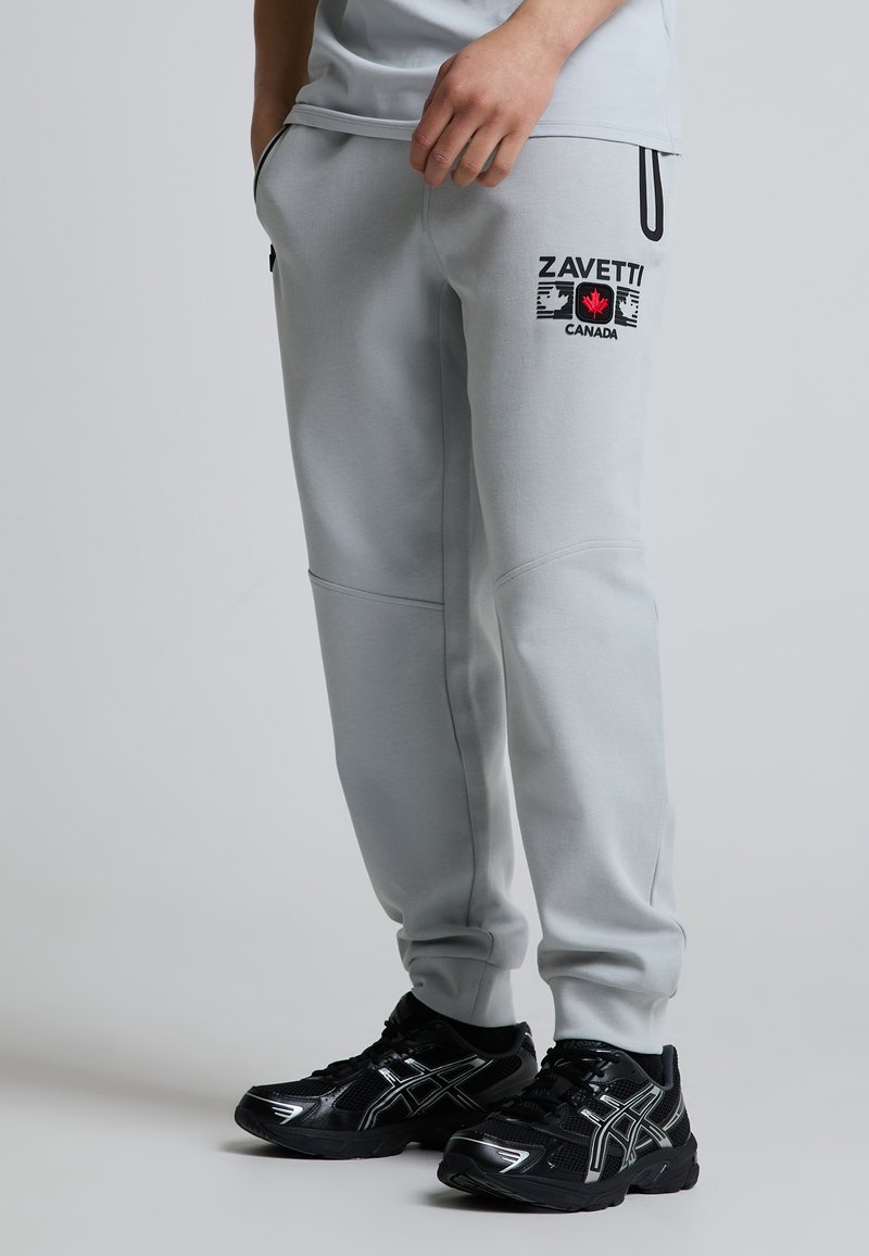 Person iført lysegrå joggingbukser med "Zavetti Canada" logo og sorte sportssko med hvide detaljer, stående på en neutral baggrund.