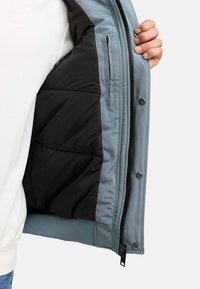 En hand som håller upp en blå jacka som visar ett svart vadderat foder, innerfickor med dragkedja och tryckknappar längs dragkedjans klaff.