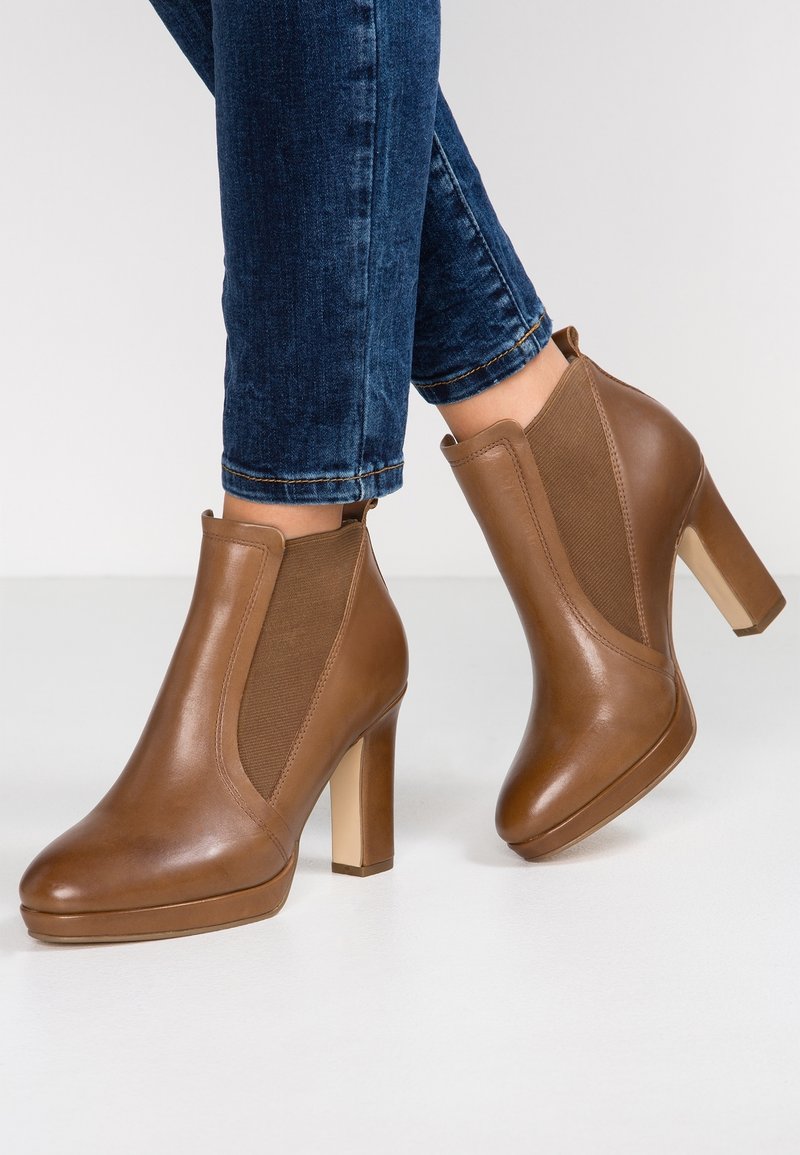 Bottes chelsea en cuir marron avec panneaux élastiques sur les côtés et talons blocs portées avec un jean en denim bleu foncé sur un sol blanc.