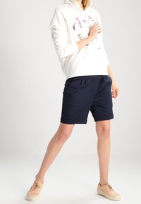 Witte hoodie met grafisch ontwerp, marineblauwe korte broek met plooien en beige sneakers, gefotografeerd tegen een neutrale achtergrond.