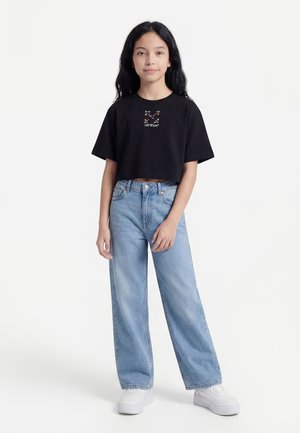 Junge Person, die ein schwarzes, kurzes T-Shirt mit Blumendesign und hellblaue, weit geschnittene Jeans trägt, steht vor einem schlichten weißen Hintergrund.