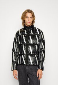 Giacca a vento nera con strisce diagonali bianche, dotata di collo alto e chiusura con zip. Tessuto liscio e lucido con sezioni trapuntate.