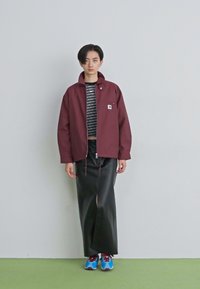 Persoon staat tegen een effen muur en draagt een bordeauxrode jas, een zwart gestreept crop top, een lange zwarte rok en felblauwe sneakers.