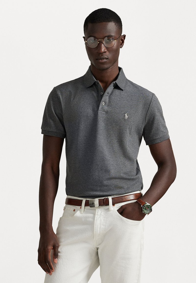 Γκρι polo μπλουζάκι με γιακά και τρία κουμπιά, με μικρό κεντημένο λογότυπο. Φορεμένο με λευκά παντελόνια και καφέ ζώνη.