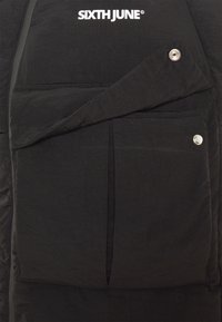 Schwarze gepolsterte Jacke mit einer horizontalen Reißverschlusstasche und einer Klappentasche mit Druckknopfverschluss. Glatte Textur und leichtes Material.