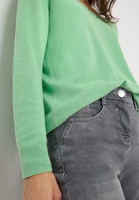 Vaaleanvihreä pehmeä neule, jossa on v-pääntie, yhdistettynä harmaan denimin farkkuihin, jotka korostavat sileää tekstuuria ja standardia viiden taskun muotoilua.