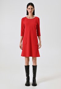 Abito rosso al ginocchio con scollatura rotonda, maniche a tre quarti e due tasche frontali; tessuto liscio e design a tinta unita.