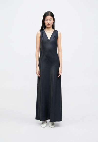 Αμάνικο, φόρεμα μέχρι το πάτωμα από σκούρο ναυτικό ύφασμα με διακριτική γυαλάδα, που διαθέτει V-λαιμόκοψη και λεπτομέρεια πλευρικής drape, συνδυασμένο με λευκά μυτερά παπούτσια.