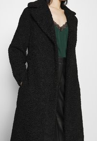 Manteau noir texturé à motif floral, doté d'un col châle et de poches ; superposé à un haut vert avec une bordure en dentelle noire.