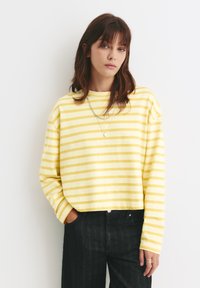 Žlutý a bílý pruhovaný dlouhý rukáv crop sweatshirt s kulatým výstřihem, spárovaný s černými džíny s vysokým pasem.