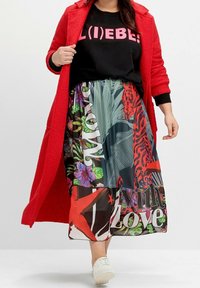 Długa, kolorowa spódnica z wyrazistymi wzorami i tekstem, wykonana z lekkiego materiału. Zestawiona z czarnym swetrem i czerwonym płaszczem. Białe buty.