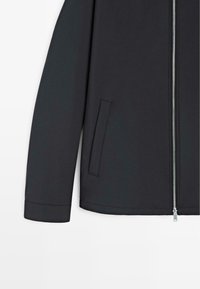 Schwarze Jacke mit langen Ärmeln, silbernem Reißverschlussverschluss und einer vertikalen Schlitztasche auf der rechten Seite.