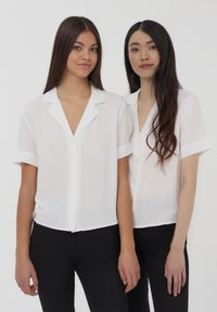 Camicie bianche a maniche corte con un taglio rilassato, dotate di colletto e polsini a contrasto, indossate con pantaloni neri.