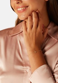 Gouden kettingscharnier met een klein kruisje, gedragen aan een hand. De blouse is van roze satijn met een knoopmanchet. Soepele, zachte textuur.