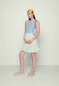 Mulher grávida com uma blusa azul sem mangas, saia floral verde clara, lenço laranja na cabeça, segurando uma carteira lilás, usando sandálias de plataforma, de pé.