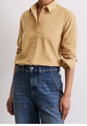 Kvinde iført beige langærmet skjorte med knapper og ærmerne rullet op samt høj talje blå denimjeans med forlommer.