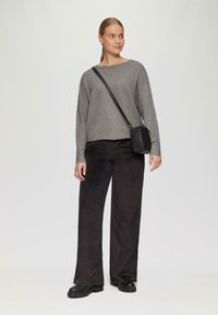 Grau gerippter Strickpullover, weite schwarze Cordhosen, schwarze Umhängetasche. Modell steht in entspannter Haltung. Schuhwerk: schwarze Plateauschuhe.