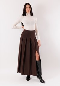 Donna con lunghi capelli scuri che indossa un dolcevita bianco, gonna plissettata marrone scuro con spacco laterale e stivali neri alti fino al ginocchio con punta appuntita.