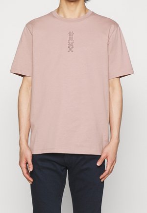 Osoba ubrana w jasnofioletową koszulkę z minimalnym pionowym czarnym abstrakcyjnym wzorem na środku klatki piersiowej, zestawioną z ciemnymi spodniami.