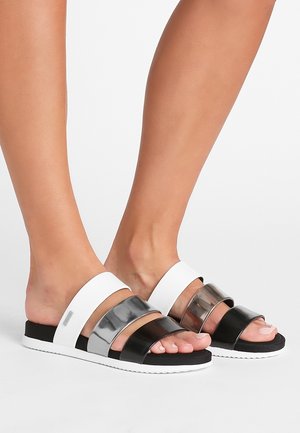 Stopy w czarnych, srebrnych i białych sandałach wsuwanych z trzema szerokimi poziomymi paskami i płaską białą podeszwą na białym tle.