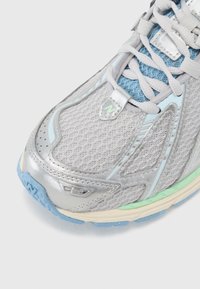 Jasnoszary but sportowy z siateczkową cholewką, srebrnymi akcentami, niebieskimi sznurowadłami oraz teksturowaną podeszwą z zielonymi i niebieskimi akcentami.