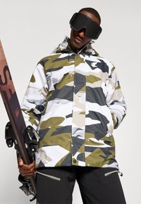 Zöld, fehér és szürke álcázott mintaű dzseki, kapucnival, cipzáras zsebekkel, fekete sínadrággal és sílécel viselve.