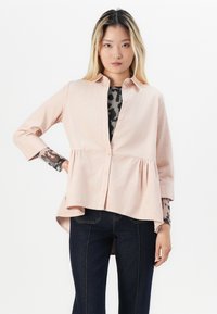 Camicia button-up rosa chiaro con orlo a peplum, realizzata in tessuto testurizzato. Maniche lunghe con polsini, indossata sopra un top a motivo nero e grigio.