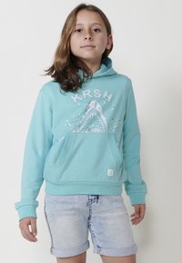 Tirkīza kapuce ar haizivs grafiku, priekšējā kabata un ribota apakšmala. Apvienota ar gaišām džinsa šortiem.