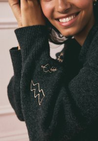 Schwarzer gestrickter Pullover mit gerippten Bündchen. Bestickte Details umfassen ein Blatt und ein Zickzackmuster in Gold. Die weiche Textur ist sichtbar.