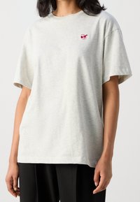 Szara bawełniana koszulka z luźnym krojem, okrągłym dekoltem i krótkimi rękawami. Zawiera czerwony haft na lewej stronie klatki piersiowej.