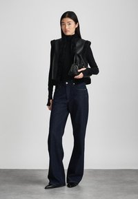 Svart faux fur- og skinnvest over en svart genser med høy hals, kombinert med mørkeblå vide jeans og en liten svart håndveske.