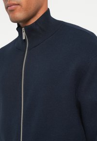 Sweater azul-marinho com fecho em zíper e colarinho canelado; apresenta um zíper prateado na parte da frente para fecho. Vista de perto.