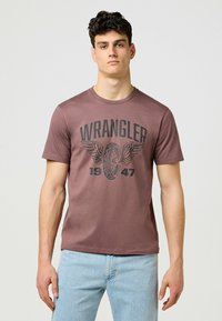 Brun kortärmad t-shirt gjord av mjukt tyg med en stor svart "WRANGLER" logotyp och vingdesign med "1947" under.
