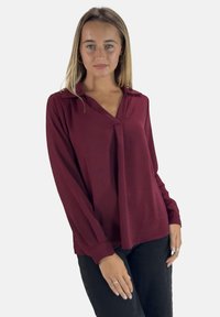 Bordeaux langharige blouse met een V-hals en subtiele textuur, met een losse pasvorm en knoopaccenten bij de manchetten. Gedragen met zwarte broeken.