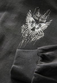 Tumši pelēks krekls ar baltu grafiku, kurā attēlots tauriņš ar spārniem un izsmalcinātu tekstu, ribota aproce un mīksta auduma tekstūra.