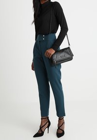 Žena oblečená v teal vysokých kalhotách s černým žebrovaným topem, černých špičatých vysokých podpatcích a nosí černou koženou kabelku přes rameno.