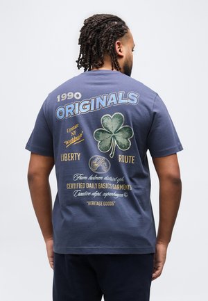 Mężczyzna z zaplecionymi włosami, ubrany w niebieską koszulkę z dużym zielonym koniczyną i tekstem zawierającym "1990 Originals", "Liberty Route" oraz innym nadrukiem z tyłu.