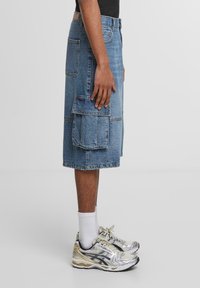 Falda cargo de mezclilla en azul claro, hasta la rodilla, con múltiples bolsillos grandes. Combinada con zapatillas deportivas grises y doradas y calcetines blancos.
