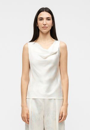 Žena s dlhými tmavými vlasmi, oblečená v bielom saténovom topu bez rukávov a svetlých širokých nohaviciach, stojaca pred jednotným pozadím.