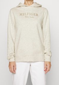 Světle béžová mikina s klokánkovou kapsou, stahovací kapucí a zlatým logem "HILFIGER New York". Měkká textura a dlouhé rukávy.