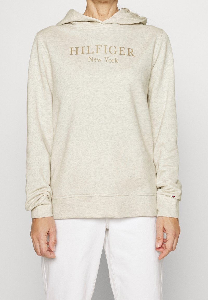 Světle béžová mikina s klokánkovou kapsou, stahovací kapucí a zlatým logem "HILFIGER New York". Měkká textura a dlouhé rukávy.