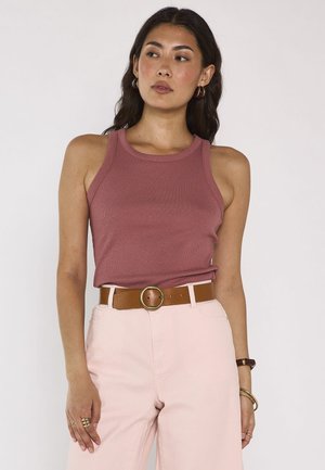 Woman wearing a sleeveless dusty rose tank top, light pink high-waisted pants with a brown belt, and gold hoop earrings.