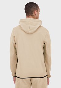 Sudadera con capucha beige de textura suave, con cordón en la capucha y banda negra en el dobladillo, vista desde la parte trasera.