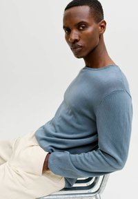 Homme aux cheveux courts et portant une boucle d'oreille, vêtu d'une chemise à manches longues bleue et d'un pantalon beige, assis de côté sur une chaise en métal.