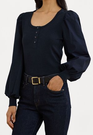 Vrouw draagt een aansluitende zwarte lange mouwen blouse met opgeblazen mouwen en een met knopen gesloten hals, ingestopt in donkere jeans met een zwarte riem.