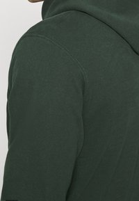 Primer plano de una persona que lleva una sudadera con capucha de color verde oscuro, mostrando el área del hombro y el cuello con detalles de costura visibles.