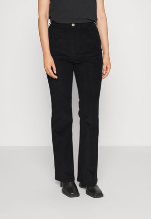 Personne portant un pantalon noir taille haute à jambes droites, un haut noir et des chaussures noires à talons, debout devant un fond clair uni.