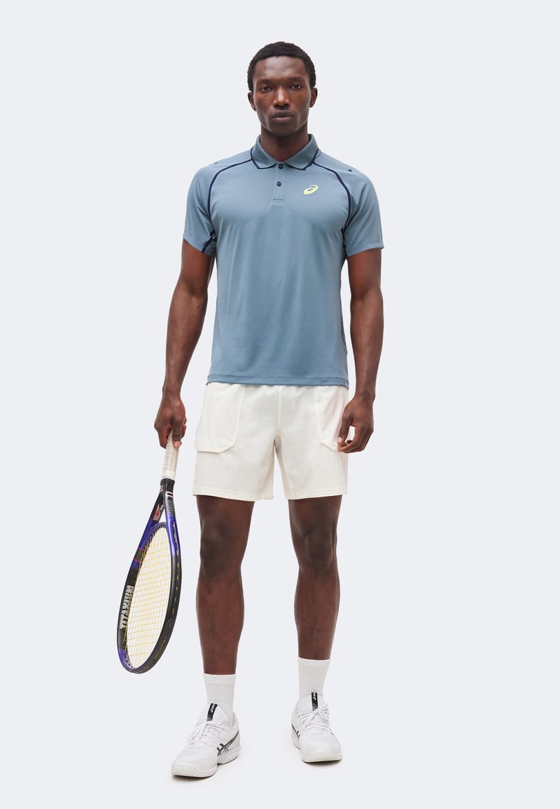 Mann steht in blauem Poloshirt und weißen Shorts, hält einen Tennisschläger, trägt weiße Socken und Sportschuhe vor einfarbigem Hintergrund.